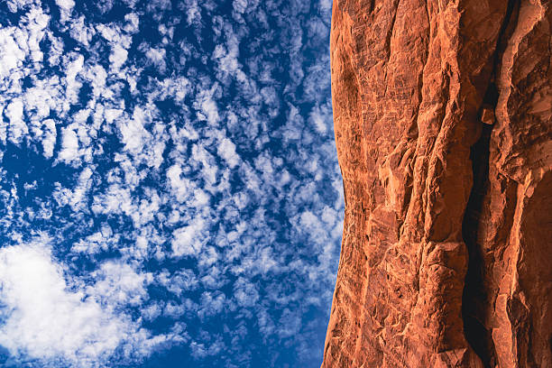 Close-up view of a dramatic sandstone rock formation in Arches National Park, Utah, set against a vivid blue sky filled with cirrocumulus clouds. The vertical composition highlights the natural textures, erosion patterns, and raw beauty of the American Southwest desert landscape.