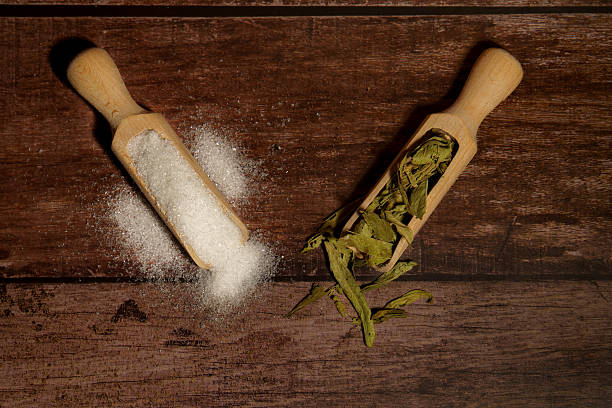 Scoops with natural stevia extract leaves and white granulated sugar on a dark wooden background, showing healthy alternatives
