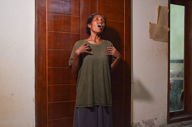 A woman in a green shirt stands against a dark wooden door with a shocked facial expression, gasping with her hands placed near her chest in a dramatic and surprised pose.