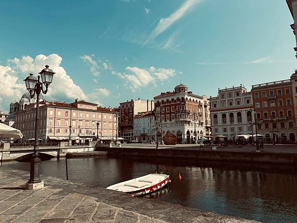 View of the Canal Grande in Trieste, Italy. Historic buildings with red and white facades line the canal. A small boat is moored in the foreground.