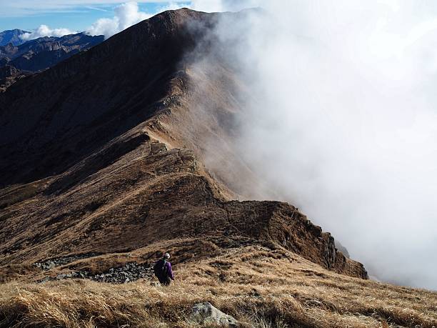 A lone trekker descends a steep, golden grassy ridge in the Northern Apennines, with thick white clouds on one side and the rocky peak of Mount Marmagna in the distance.