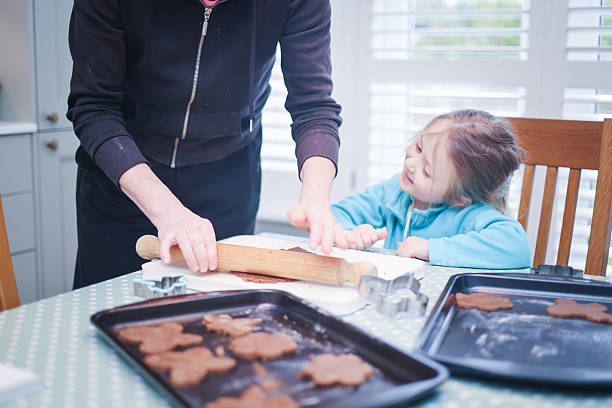 Five year old girl smiling as she looks at her Grandmother roll out gingerbread dough as they prepare to make Gingerbread shapes at a domestic kitchen table