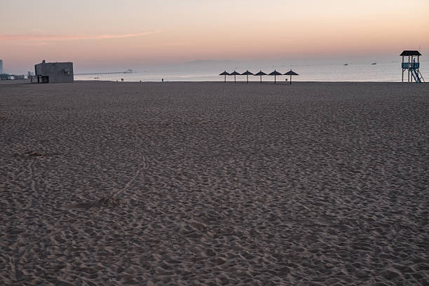 A wide panoramic view of an empty sandy beach featuring a silhouette of a lifeguard tower and closed beach umbrellas against a soft morning sky.