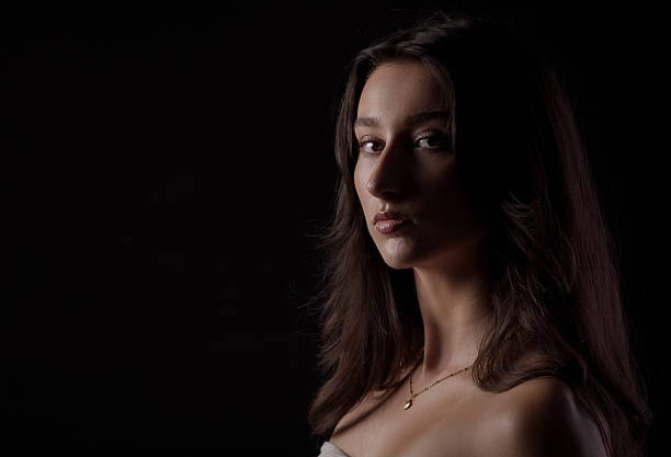 Studio portrait of a young Caucasian woman with long brown hair, wearing a delicate gold necklace, illuminated by soft lighting against a dark backdrop