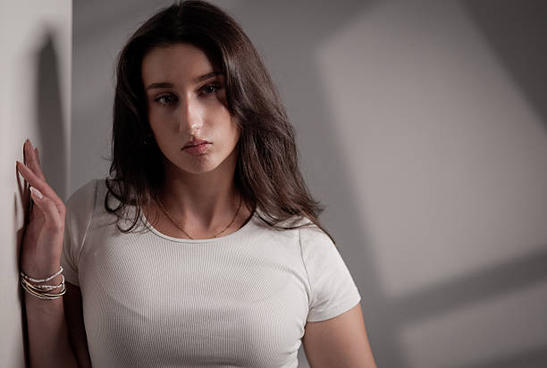 Sad young woman with long brown hair and white t shirt leaning against white wall with dramatic shadows