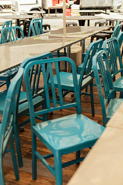 Chairs are lined up around several tables in a market setting. The chairs are painted blue, and the tables have a flat surface. The scene shows no people present