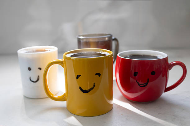 Four mugs with smiley faces are placed on a table. Each mug is filled with hot coffee. The sunlight shines on the table, highlighting the cheerful colors of the mugs and their contents.