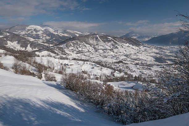 Wide panoramic view of a snowy rural mountain landscape in winter. Rolling hills, scattered village houses and forested slopes covered in snow under a dramatic sky. Tranquil winter scenery ideal for concepts of rural life, nature, winter travel, countryside tourism, climate and seasonal lifestyle.