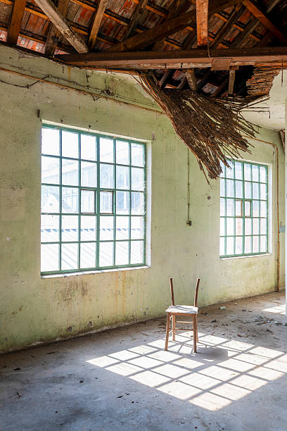 A single wooden chair stands in an abandoned school classroom. Sunlight streams through a large grid window, casting strong geometric shadows on the dusty floor. Peeling walls and scattered debris create a quiet, melancholic atmosphere of abandonment and forgotten education.