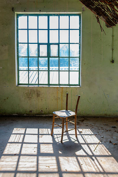 A single wooden chair stands in an abandoned school classroom. Sunlight streams through a large grid window, casting strong geometric shadows on the dusty floor. Peeling walls and scattered debris create a quiet, melancholic atmosphere of abandonment and forgotten education.