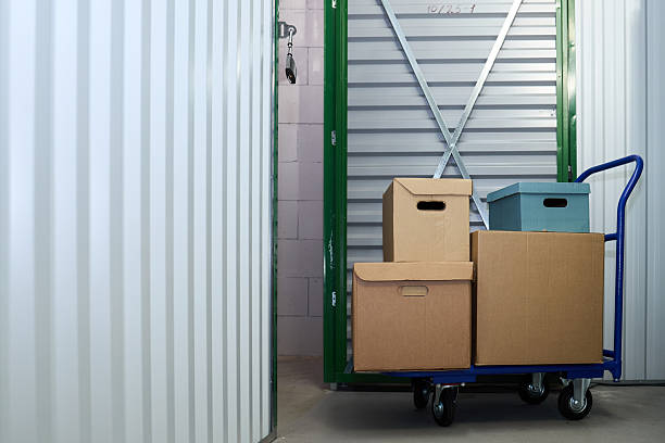 Stacked cardboard boxes on blue hand truck standing in front of open storage unit door, showing organized moving or storage process in self storage facility interior