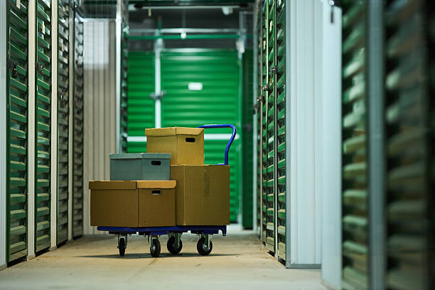 Stack of cardboard boxes on blue hand truck standing in corridor of self storage facility, green metal storage unit doors lining both sides, concrete floor visible, no people present