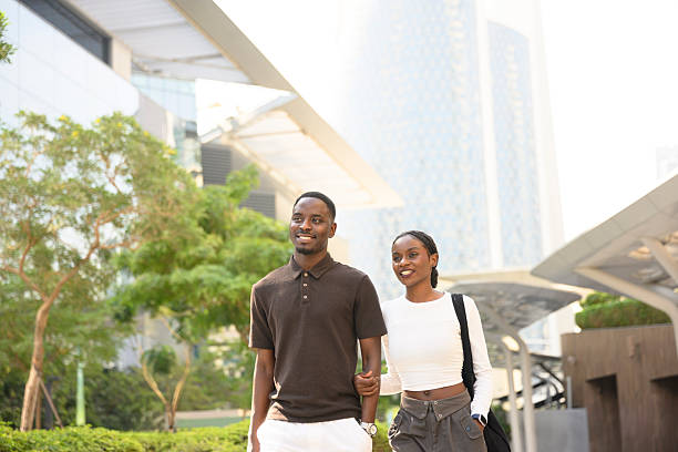 Young couple walking arm in arm through a modern city park, smiling and enjoying a carefree, warm afternoon together amid trees, architecture and urban greenery