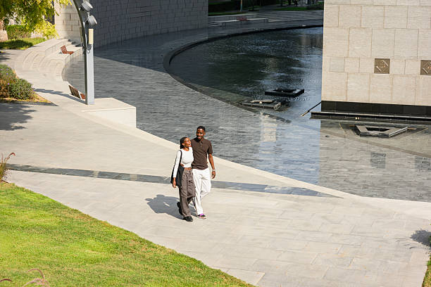Couple walking hand in hand along a modern stone pathway outdoors next to a contemporary water feature, enjoying a leisurely afternoon exploring architecturally significant grounds