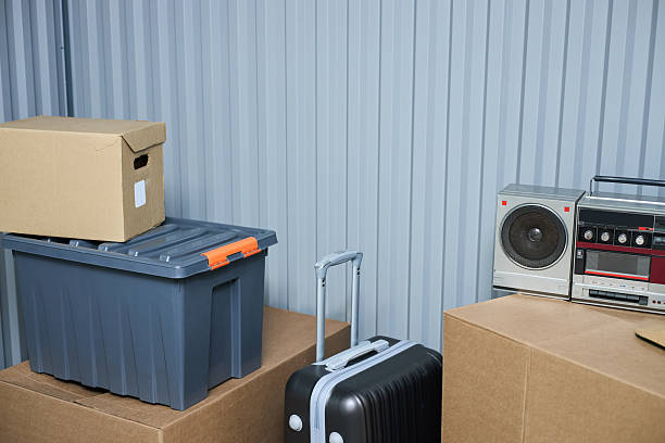 Stacked cardboard boxes, plastic storage container, hard shell suitcase with extended handle, vintage radio and cassette player arranged against corrugated metal wall in storage unit