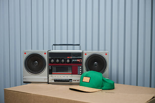 Vintage boombox stereo sitting on cardboard box with green baseball cap in foreground, retro audio equipment and casual accessory arranged against striped wall background