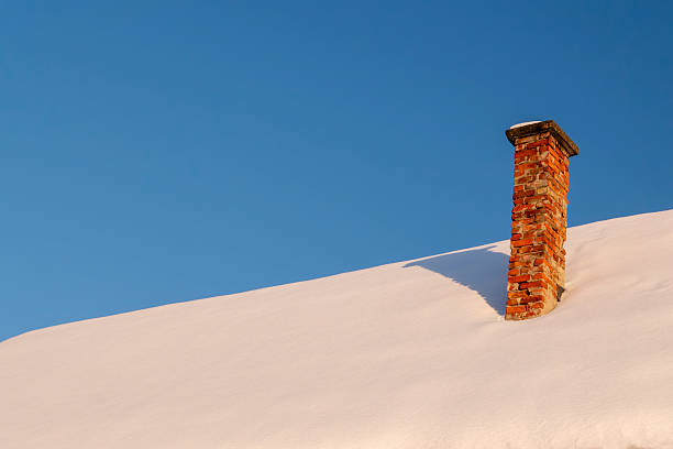 Minimal winter scene showing a brick chimney rising from a snow covered roof under a clear blue sky. Conceptual image representing winter, isolation, warmth, home, shelter, simplicity, and contrast between cold and warmth. Ideal for backgrounds and copy space.