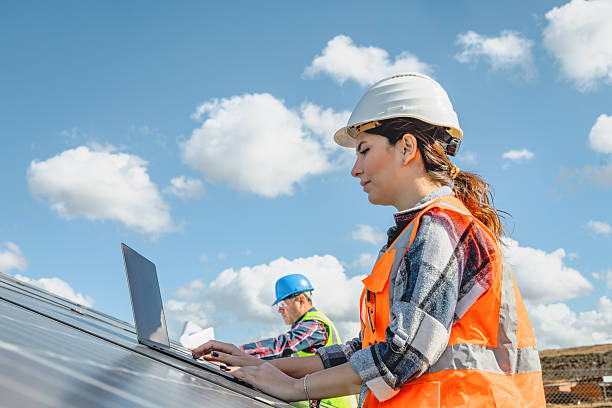A female renewable energy engineer and a male solar technician wearing safety helmets and high visibility vests work together while checking solar panels at an outdoor photovoltaic power station, the engineer uses a laptop for system analysis while the technician performs technical inspection, clean energy production, sustainable technology, teamwork, engineering, maintenance, and modern renewable energy industry concept, isolated natural background with blue sky and clouds