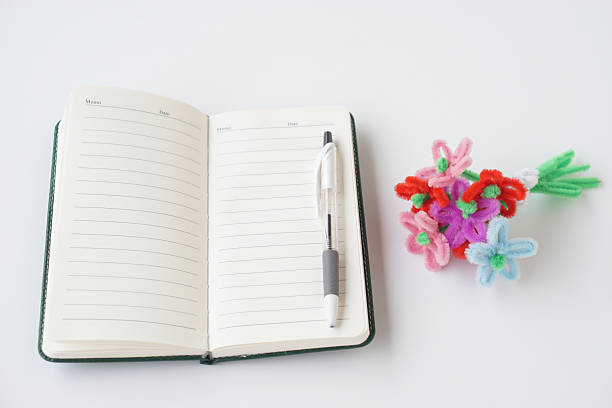 Open blank notebook with pen placed on white background next to colorful handmade craft flowers. Creative workspace concept for planning, writing, education, DIY art, imagination and inspiration