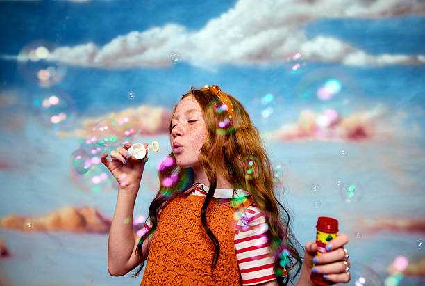 Young girl with red hair blowing colorful soap bubbles against a painted sky background