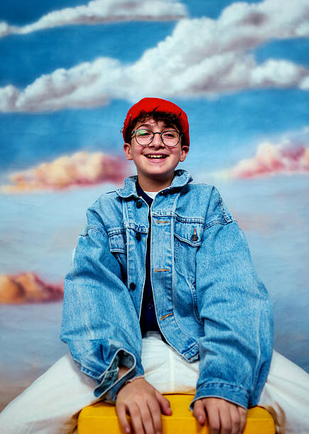 Young boy smiling, wearing glasses and a denim jacket, in front of a cloud backdrop
