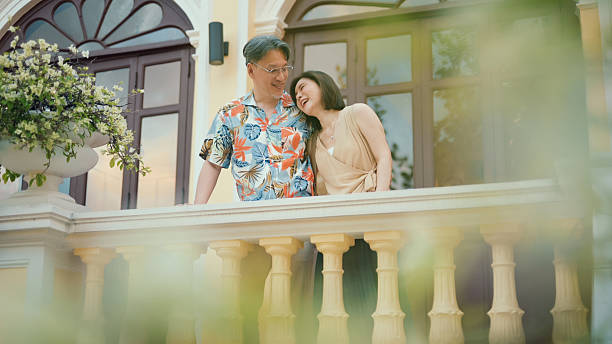 senior asian couple standing on a balcony inside a boutique hotel, enjoying architectural scenery and quiet moments together. the scene reflects active aging, emotional companionship, slow travel mindset, and mature lifestyle choices focused on meaningful shared experiences rather than fast tourism.