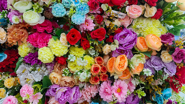 A dense and colorful top-down view of various artificial flowers, including roses, peonies, and daisies in shades of pink, red, yellow, and blue. Perfect for festive backgrounds, wedding decor concepts, or floral patterns.