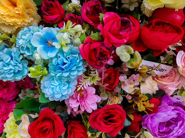 A dense and colorful top-down view of various artificial flowers, including roses, peonies, and daisies in shades of pink, red, yellow, and blue. Perfect for festive backgrounds, wedding decor concepts, or floral patterns.