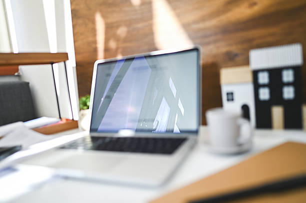 Laptop displaying reflections on screen, a coffee mug, and miniature houses on a blurred desk