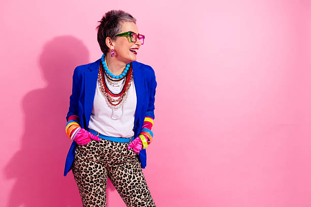A stylish mature woman with short grey hair wearing a blue blazer leopard print pants and colorful jewelry stands confidently against a pink background perfect for fashion lifestyle and advertising themes.