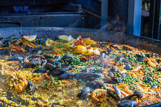 Close up of traditional Spanish seafood paella cooking in a large pan. Authentic rice dish with mussels, shrimp, and lemon wedges at a street food market. London, UK, 29 March 2024
