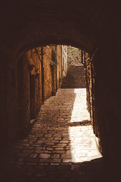 Sunlight filters through an arched alleyway, casting shadows on the cobblestone path and leading to stone steps. Italy Tuscany