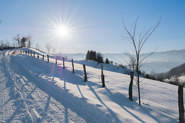 Snow covered rural hillside with wooden fence and barbed wire illuminated by low winter sun. Peaceful countryside landscape with long shadows, blue sky, and distant mountains. Ideal for concepts of freedom, calm, sustainability, rural life, seasonal weather, and winter travel backgrounds.