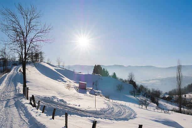 Snow covered rural hillside with wooden fence and barbed wire illuminated by low winter sun. Peaceful countryside landscape with long shadows, blue sky, and distant mountains. Ideal for concepts of freedom, calm, sustainability, rural life, seasonal weather, and winter travel backgrounds.