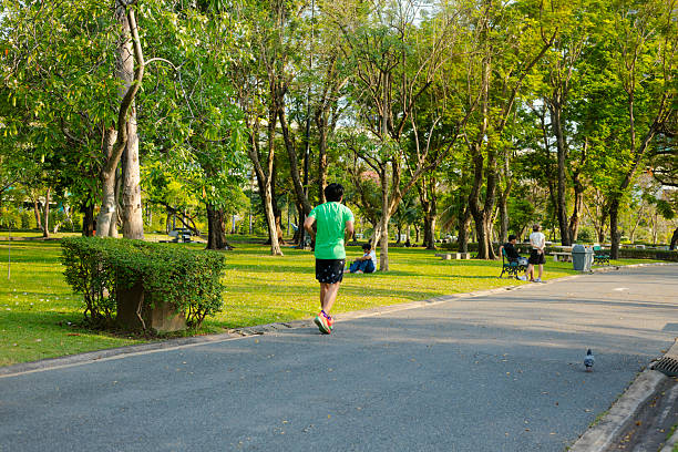 Rear view of jogging mature thai man captured on pathway in Chauchak Park in Bangkok at afternoon. Man is wearing sprts clothes and running shoes. Scene is in northern part of the park. In background are som eother thai people.  Some young people are sitting under trees. At pathway a young couple is at a bench