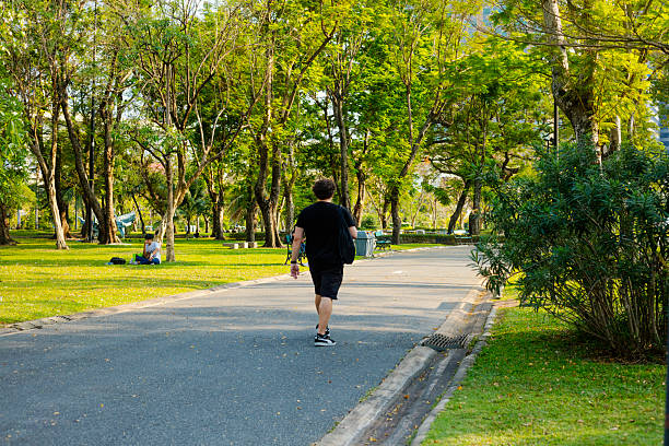 Young white man in shorts is walking in Chatuchak Park in Bangkok at afternoon. Man is wearing casual black colored clothes and he has bag around shoulder. Rear view of walking man on pathway around a lake. In background are several benches along the pathway. Behind man a person is on a bench. In background are trees