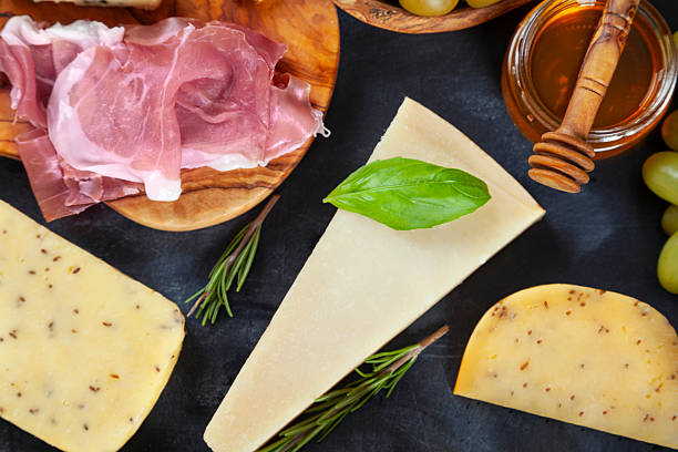 Different types of cheese, rosemary, basil, olive, honey and grapes on dark textured table, flat lay