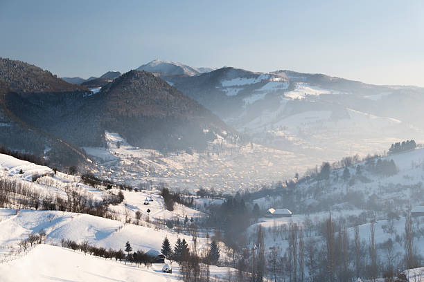 Wide panoramic view of a snow-covered mountain village and rolling hills in winter. Rural countryside landscape with scattered houses, bare trees, and soft natural light, conveying tranquility, cold season, nature, and traditional mountain life.