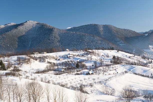 Wide panoramic view of a snow-covered mountain village and rolling hills in winter. Rural countryside landscape with scattered houses, bare trees, and soft natural light, conveying tranquility, cold season, nature, and traditional mountain life.