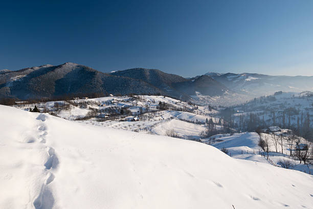 Wide panoramic view of a snow-covered mountain village and rolling hills in winter. Rural countryside landscape with scattered houses, bare trees, and soft natural light, conveying tranquility, cold season, nature, and traditional mountain life.