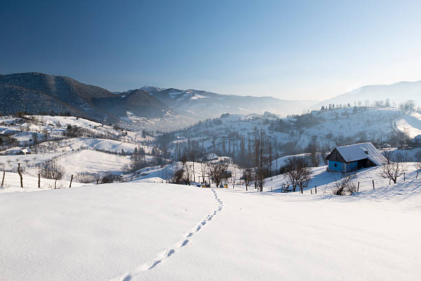 Wide panoramic view of a snow-covered mountain village and rolling hills in winter. Rural countryside landscape with scattered houses, bare trees, and soft natural light, conveying tranquility, cold season, nature, and traditional mountain life.