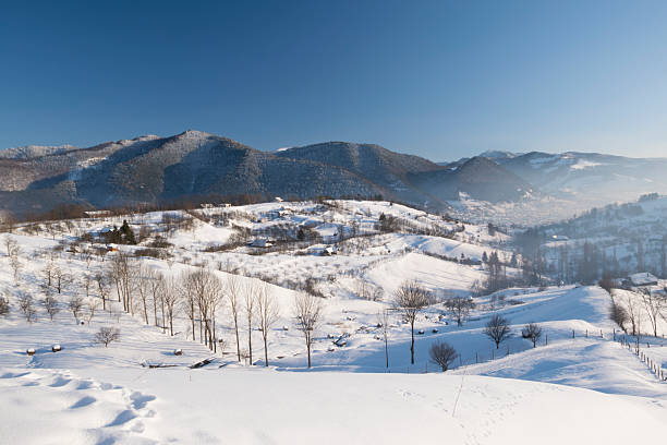 Wide panoramic view of a snow-covered mountain village and rolling hills in winter. Rural countryside landscape with scattered houses, bare trees, and soft natural light, conveying tranquility, cold season, nature, and traditional mountain life.