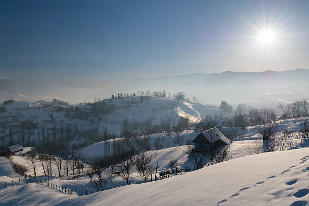 Wide panoramic view of a snow-covered mountain village and rolling hills in winter. Rural countryside landscape with scattered houses, bare trees, and soft natural light, conveying tranquility, cold season, nature, and traditional mountain life.