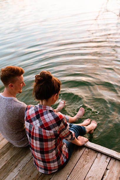 Playful young couple sitting on a wooden dock, splashing lake water with their bare feet during a relaxed summer evening.