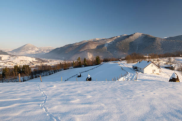 Minimalist winter landscape with human footprints crossing a snow-covered hillside, leading toward distant mountains. Clean snowy scenery with blue sky, conveying solitude, journey, freedom, exploration, and cold season nature.