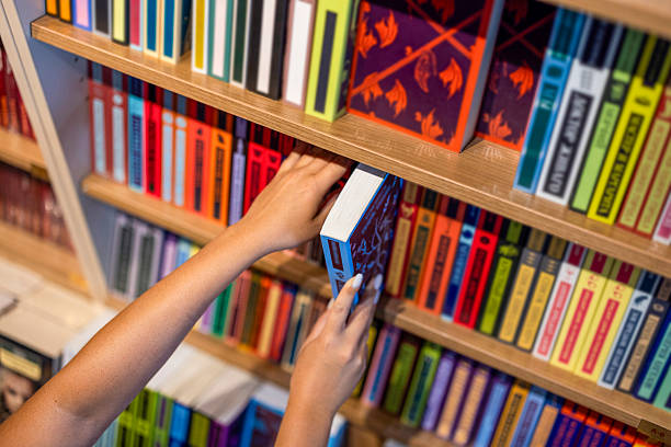 Close-up view of a young woman's hands reaching out and pulling a book from a crowded wooden bookshelf in a library or bookstore. High angle shot focusing on the selection process. Colorful book spines are visible. Concept for education, research, finding information, and literature.