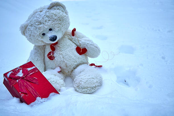 Cute teddy bear sitting in white snow with a red gift box and heart decorations. Minimal winter scene with copy space, symbol of love, Valentine’s Day, loneliness and holiday gift