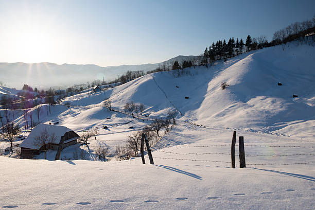 Peaceful rural winter landscape with snow covered hills, traditional haystacks, wooden fence and mountain range in the background. Scenic countryside view conveying tranquility, simplicity, sustainability, rural life and winter nature.
