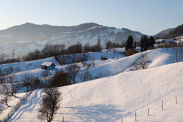 Peaceful rural winter landscape with snow covered hills, traditional haystacks, wooden fence and mountain range in the background. Scenic countryside view conveying tranquility, simplicity, sustainability, rural life and winter nature.