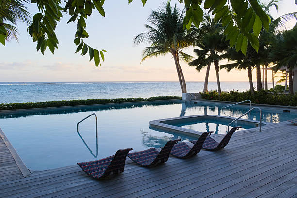 A swimming pool sits beside the ocean, framed by palm trees and coastal greenery, with calm water reflecting soft natural light. Lounge chairs line the wooden deck as the sea stretches toward the horizon, creating a peaceful coastal leisure scene.